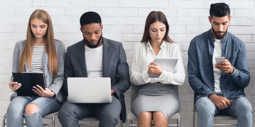 Four people seated in a row, each engaged with different digital devices, against a white brick wall.
