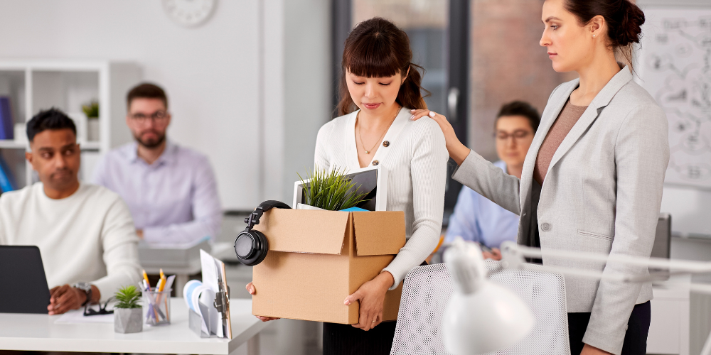 A woman carrying a box of belongings being comforted by a colleague, suggesting she may be leaving her job or transferring.