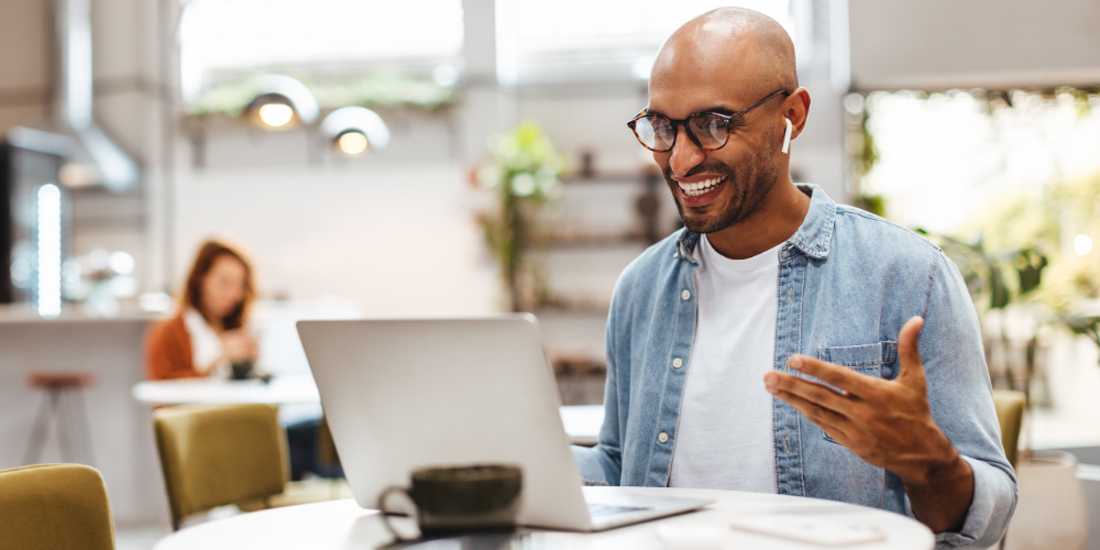 A cheerful man with glasses engaging with his laptop in a casual, modern cafe environment.