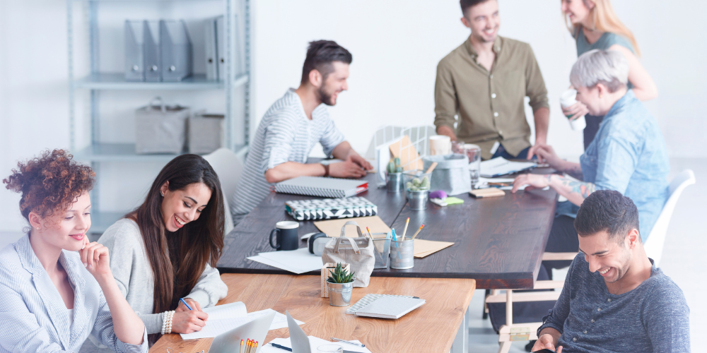 A modern office scene with a group of relaxed, smiling professionals collaboratively working at a large table.