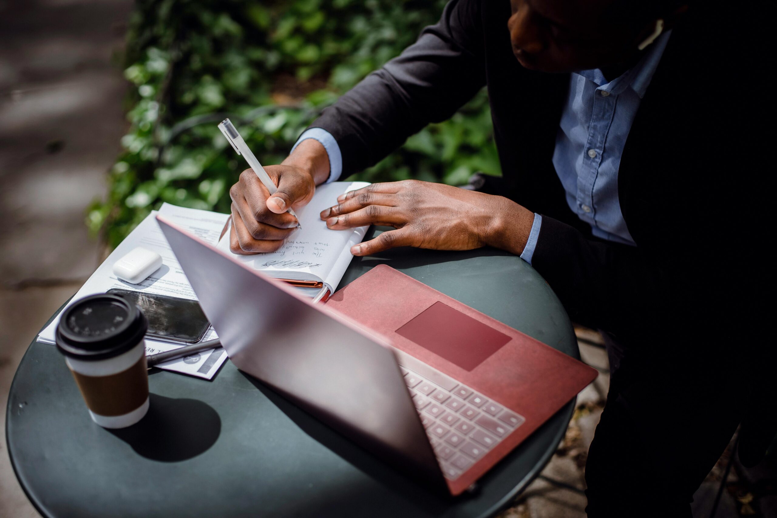 Businessman writing notes outdoors with a laptop.
