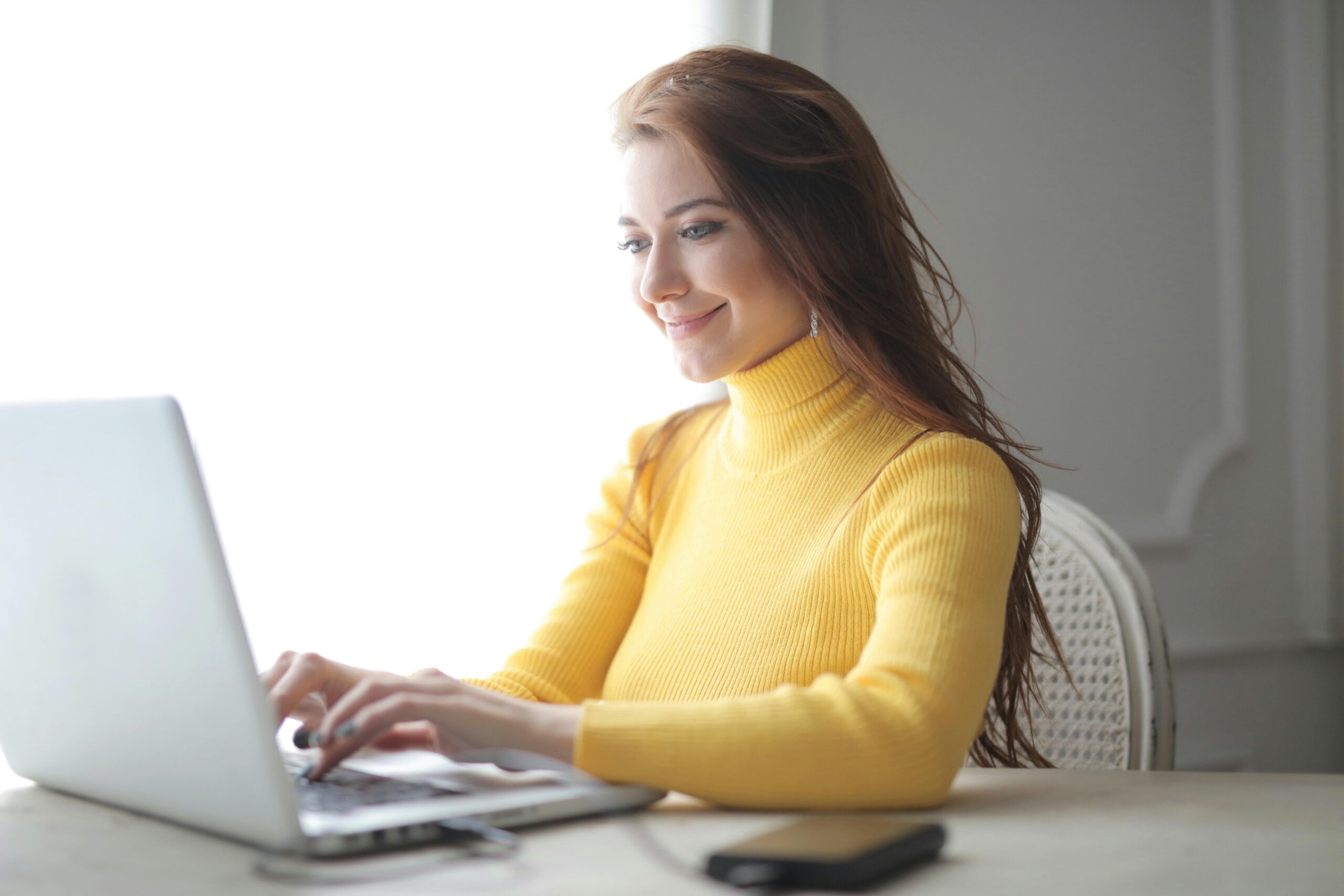 A person with obscured face wearing a yellow sweater is typing on a laptop at a white desk.