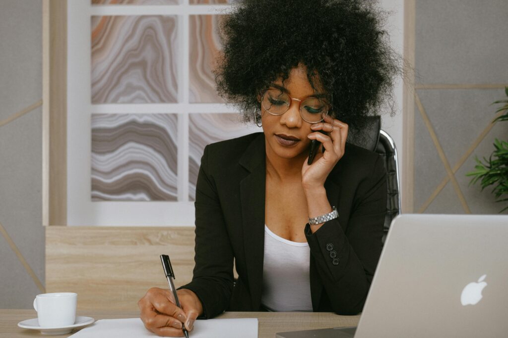 A customer support representative writing notes at a desk with a laptop and coffee in a professional workspace.