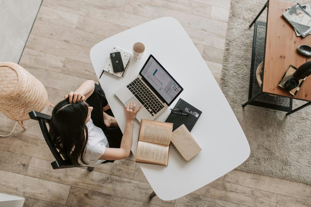 A sales rep working at a bright, white desk with a laptop, a notebook labeled "PLASSO," and various office supplies, analyzing sales performance data.