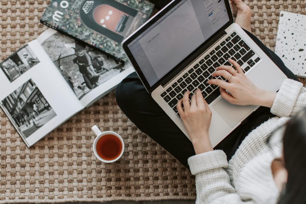 A person typing on a laptop while sitting on a carpeted floor, surrounded by open books and a cup of tea, illustrating the technical writer key skills summary.