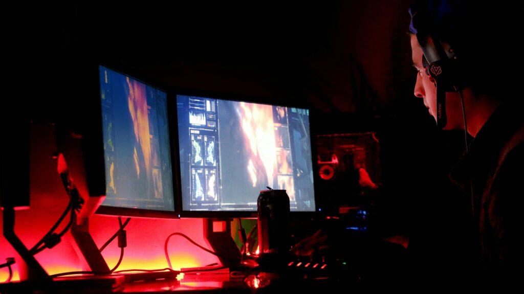 A motion graphics designer's workspace with dual monitors displaying complex visual designs and data, illuminated by red ambient lighting.