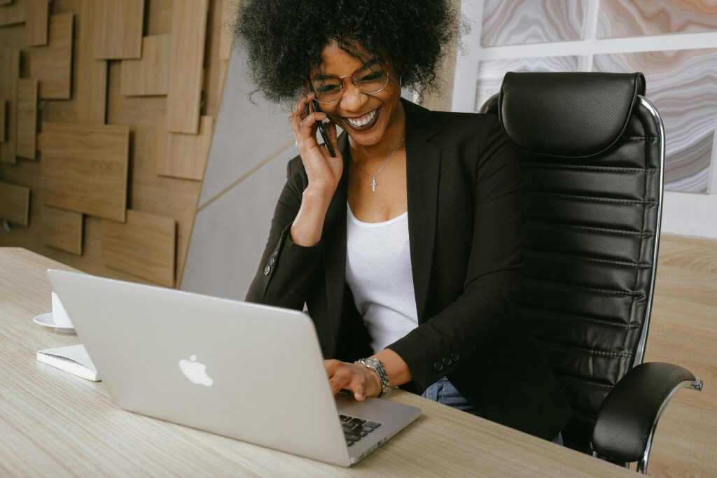 A professional woman in a black blazer and glasses sits at a desk, multitasking on her laptop while speaking on the phone, representing the essential skills of an influencer marketing manager.