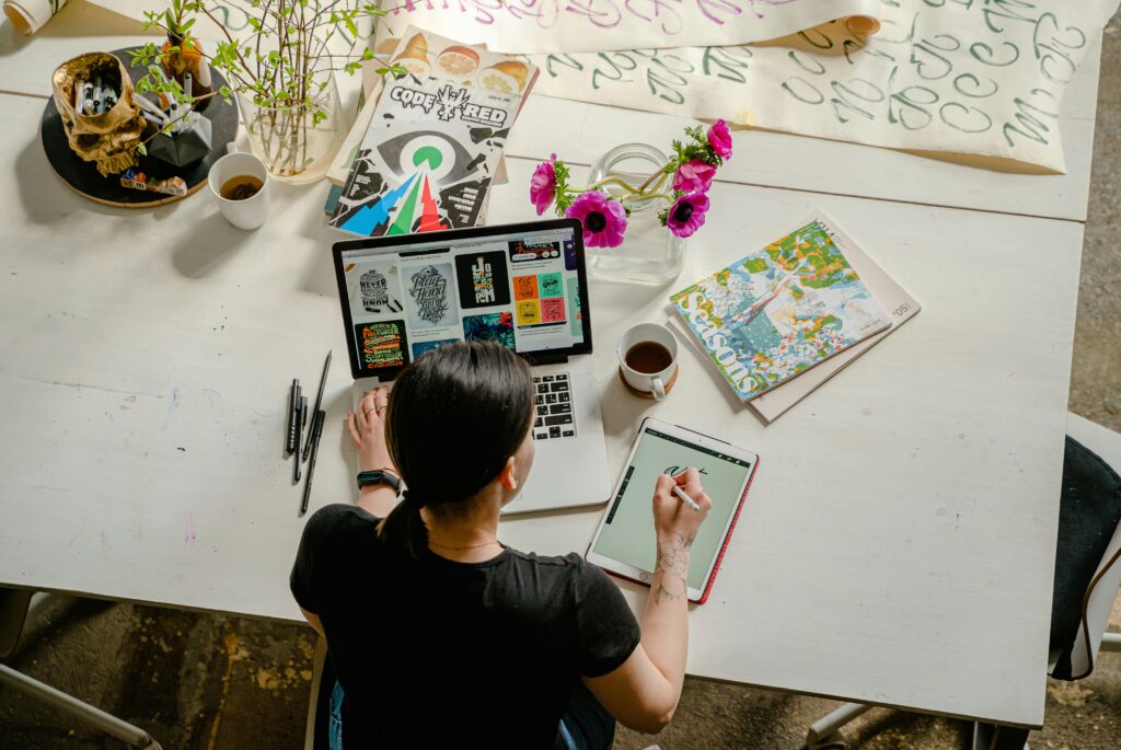 A graphic designer using a digital tablet next to a laptop on a white desk with art supplies and inspiration.