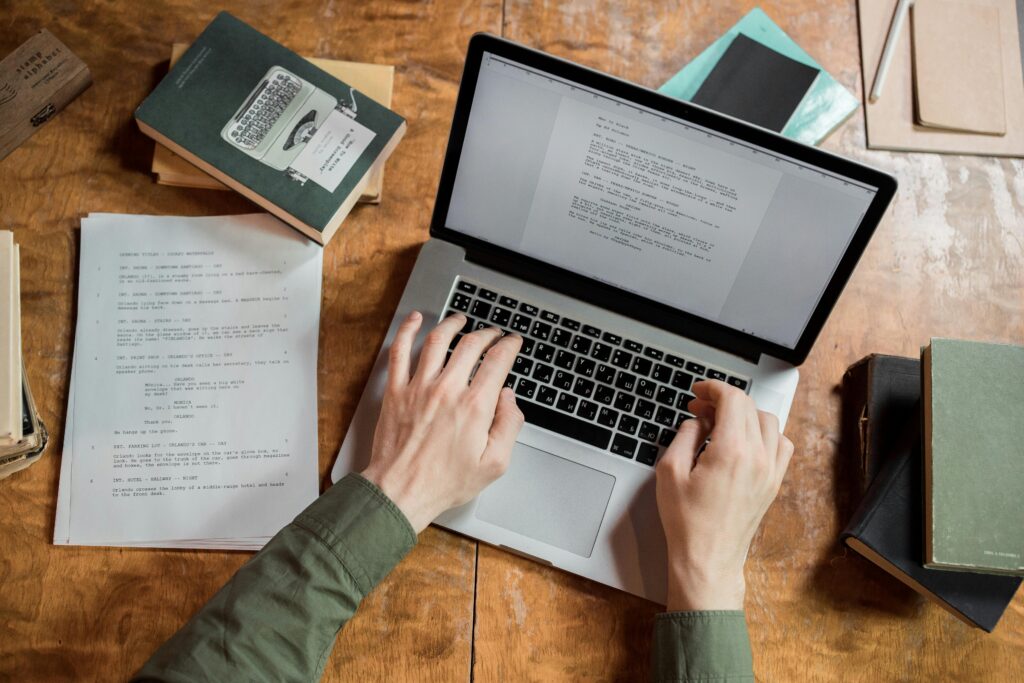A copywriter working on a laptop, typing a script while surrounded by books and notes on a wooden desk.