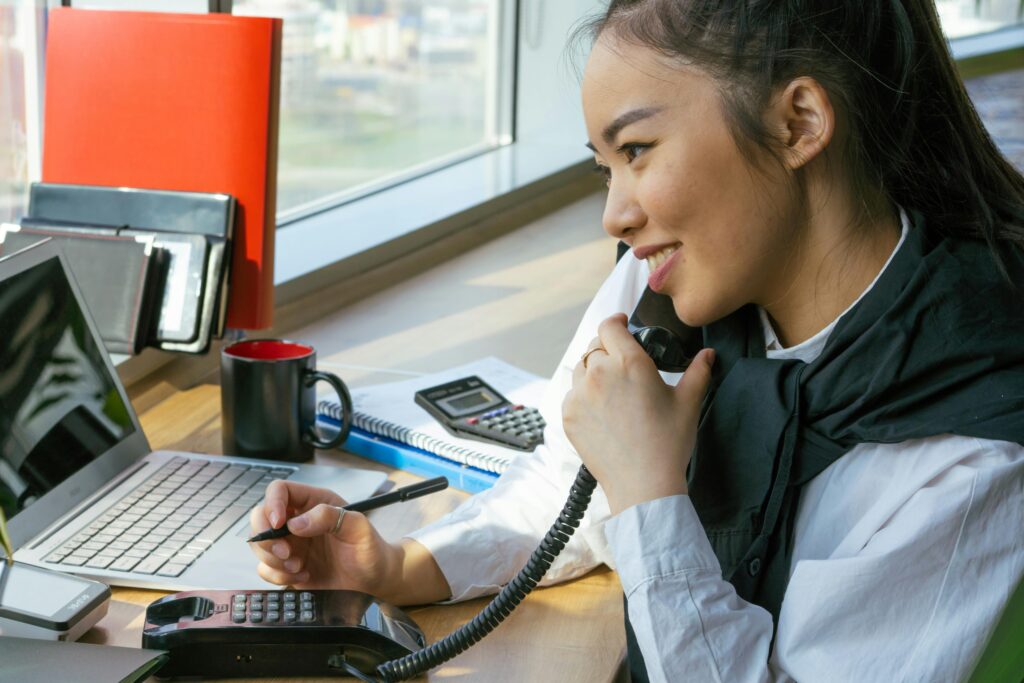 An executive assistant speaking on a corded phone while holding a pen and looking at a laptop.