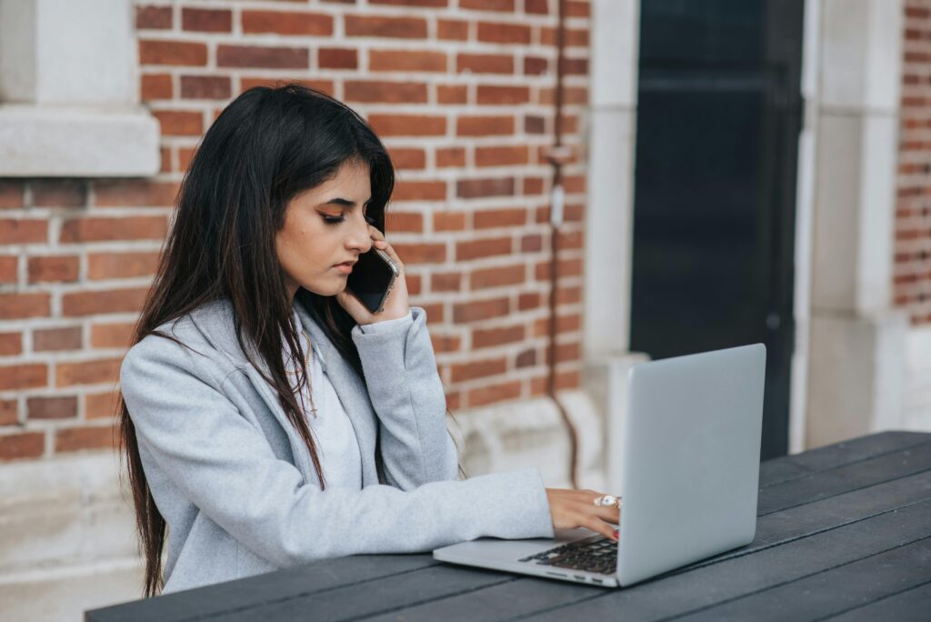 An inbound sales representative multitasking, speaking on the phone while working on a laptop at an outdoor table.