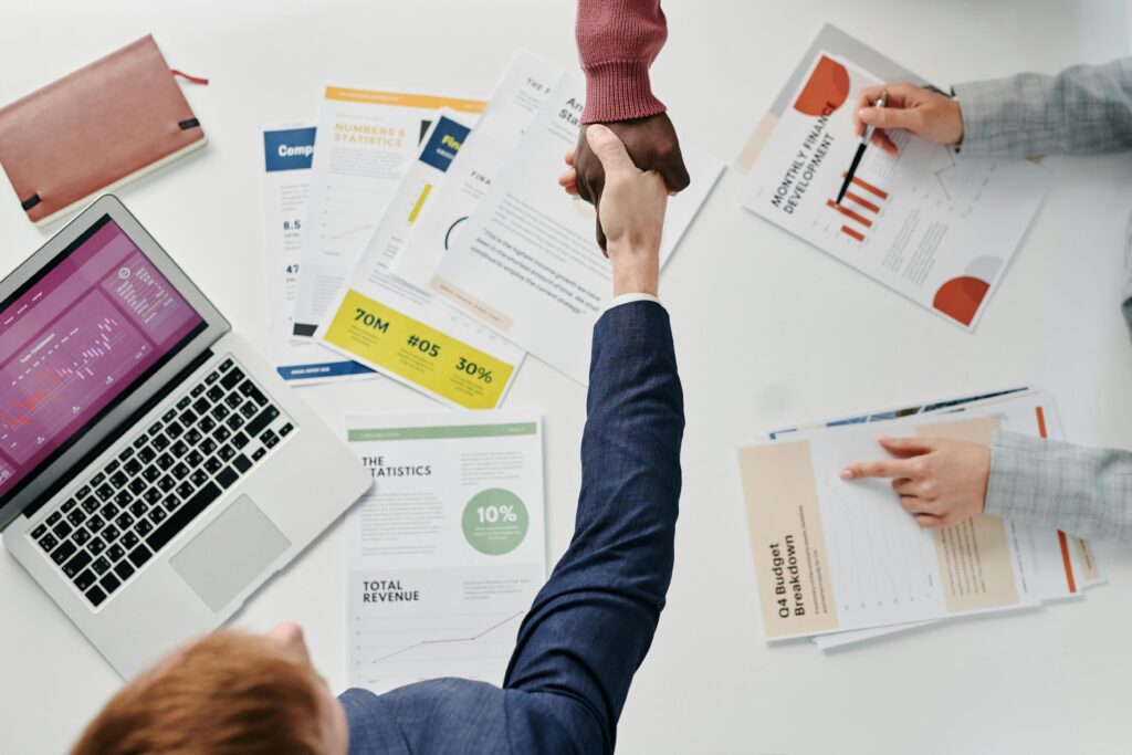 An outbound sales representative in a business suit shakes hands with a client over a desk filled with financial reports and a laptop, symbolizing a successful sales deal.