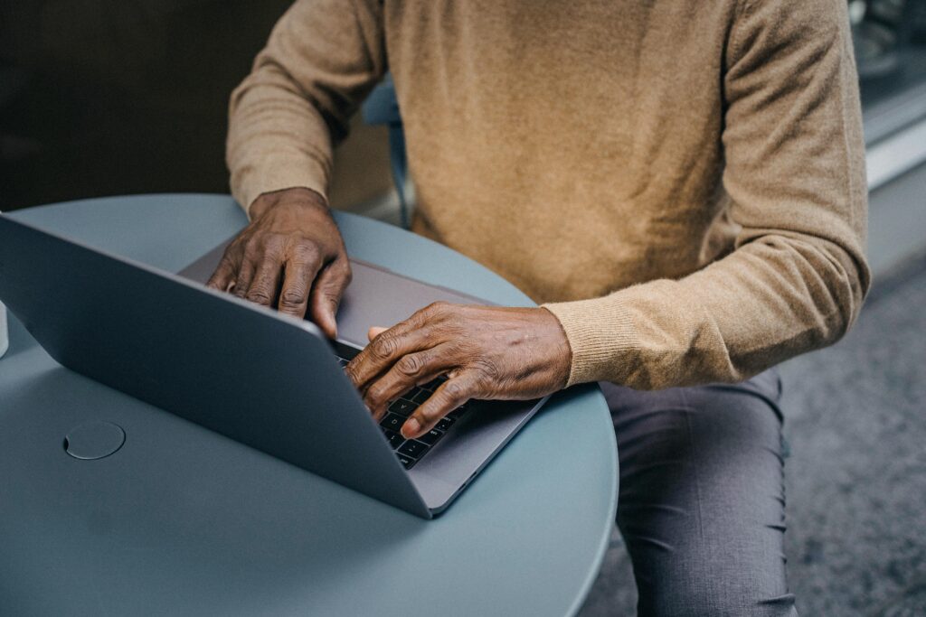 A person with dark skin, wearing a brown sweater, typing on a laptop at a small round table outdoors, suggesting remote work or a flexible work environment.