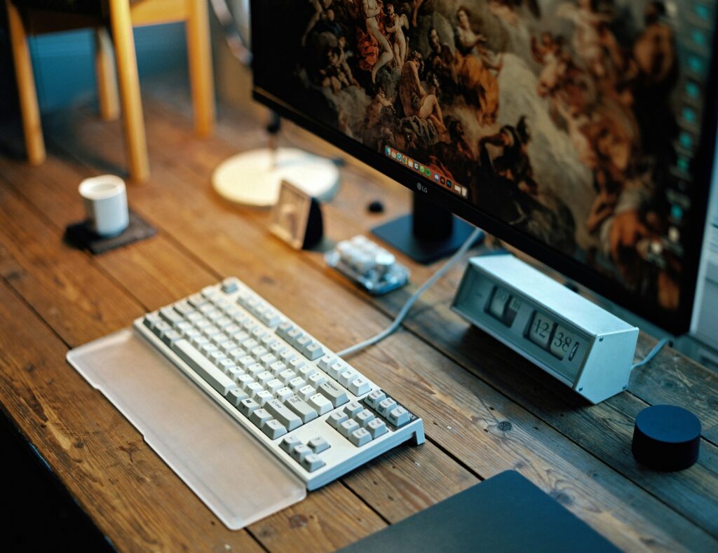 A bright home office setup with a laptop, an external monitor, a smartphone, and a water bottle on a wooden desk, positioned in front of large windows with sunlight streaming in.