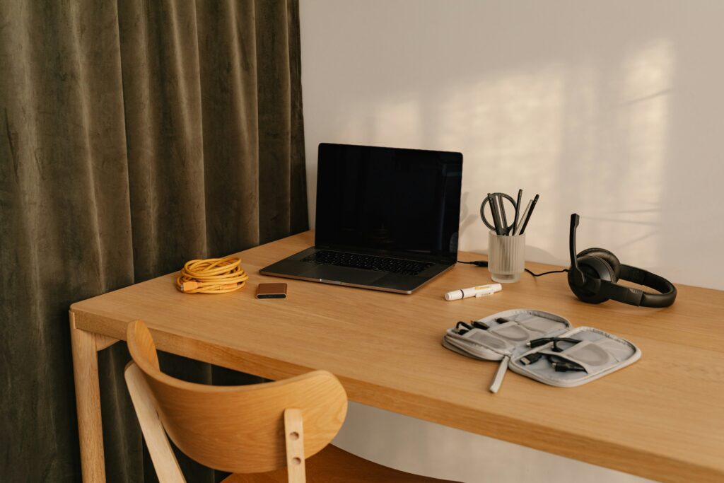 A high-angle shot of a person working on a silver laptop with a financial spreadsheet and a pie chart visible on the screen. A brown leather notebook and pen are also on the light wooden desk.