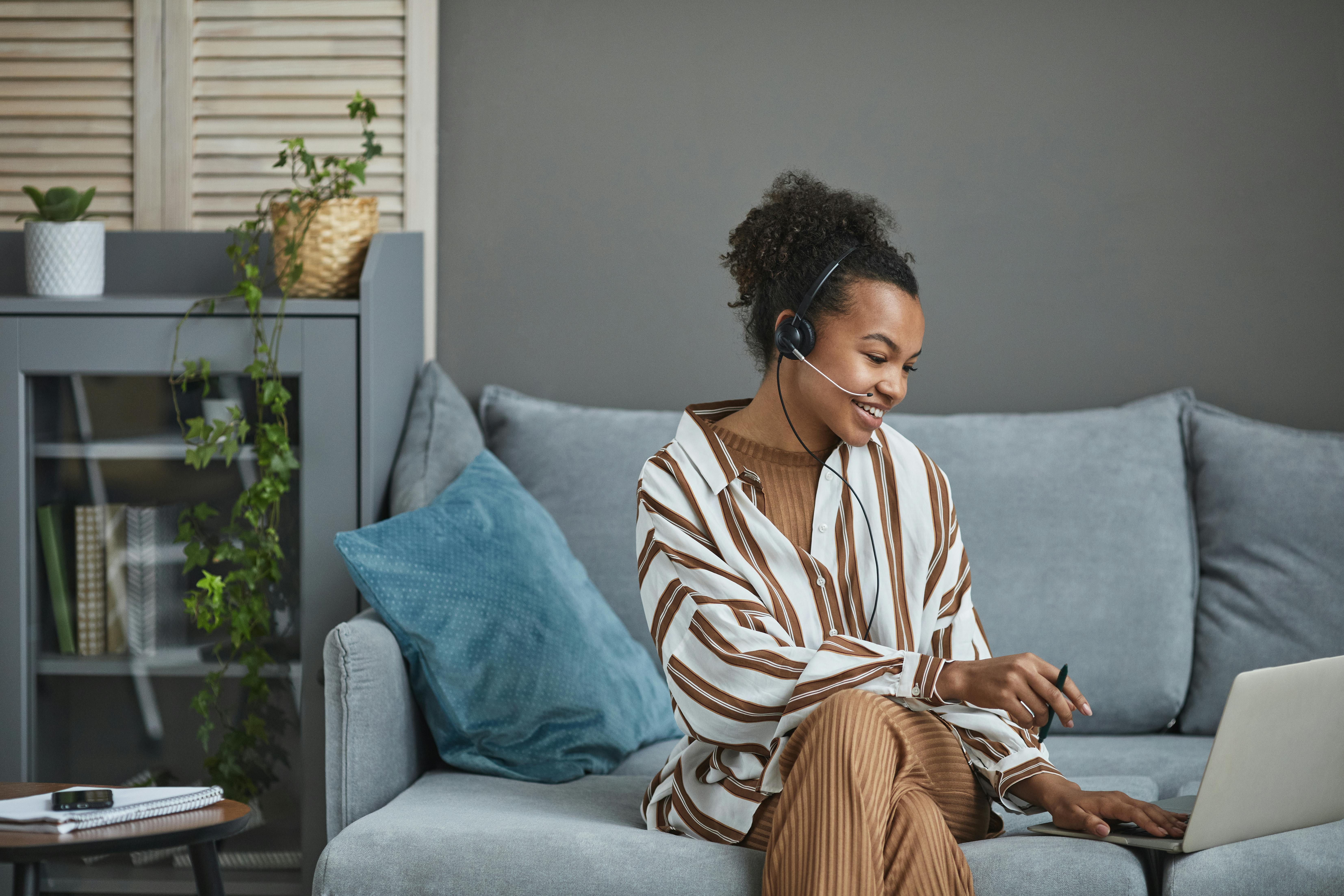 A smiling person wearing a headset with a microphone sits comfortably on a gray sofa, actively working on a laptop. This scene embodies the flexibility and professionalism of top virtual assistant services.
