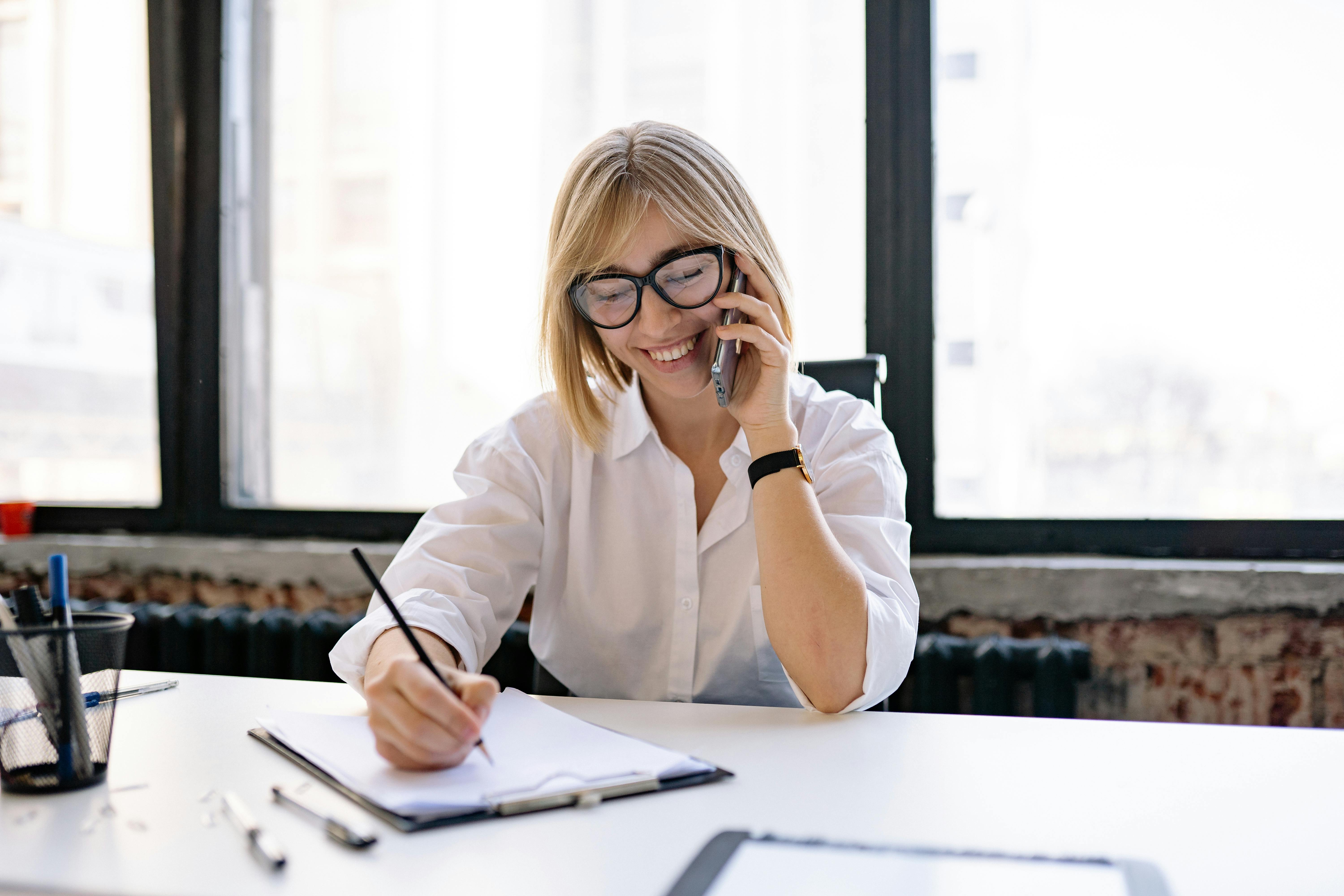 This image features a smiling woman, dressed in a crisp white shirt and stylish glasses, expertly multitasking as she talks on a mobile phone and simultaneously writes on a document on a clipboard. Her bright and engaged demeanor, set against a well-lit office background, perfectly encapsulates the energetic and communicative environment of top creative recruitment agencies. This scene highlights the constant interaction, negotiation, and detailed note-taking involved in connecting innovative talent with exciting opportunities in the creative industries.
