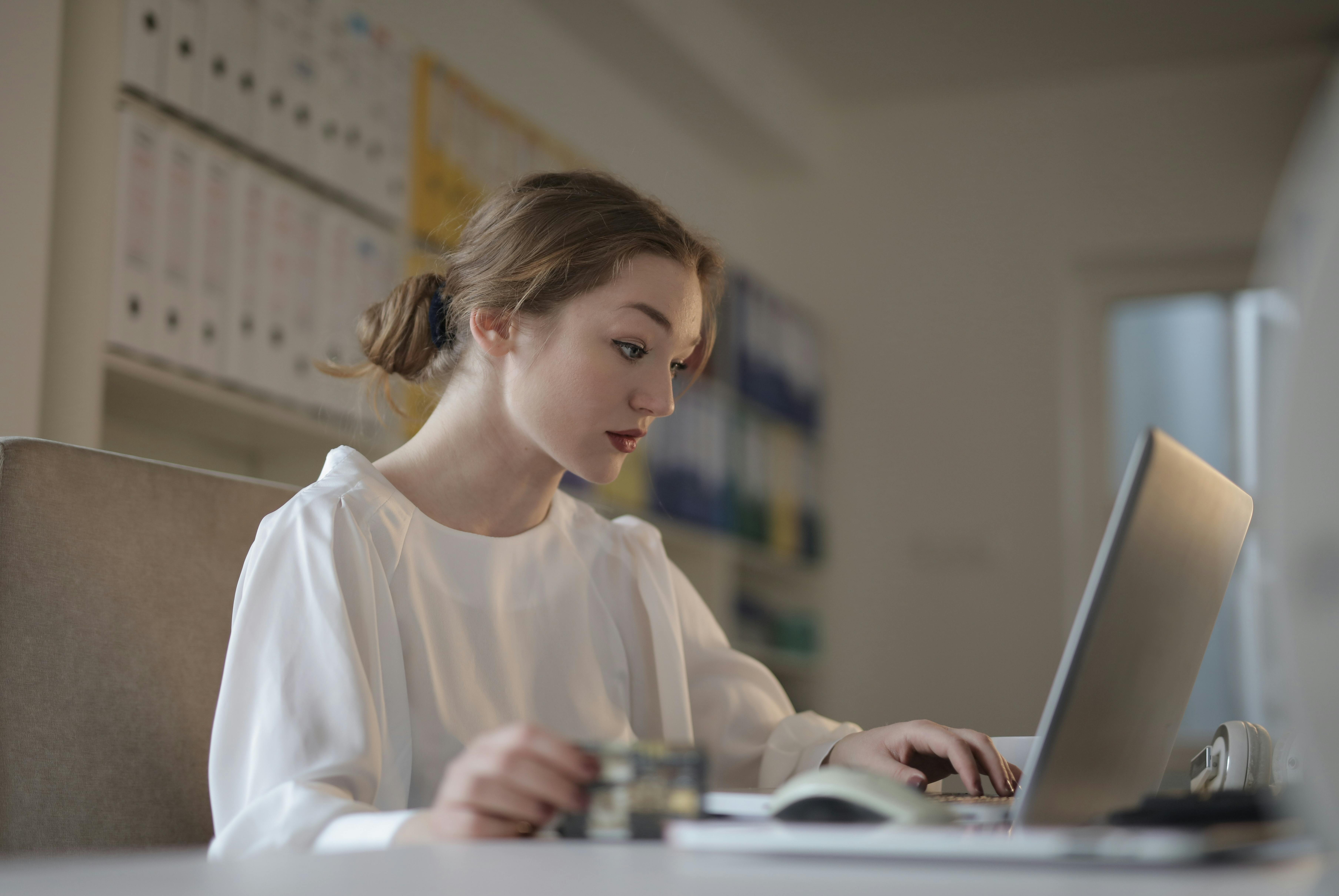A focused young woman in a white blouse is working on a laptop at a desk, holding a card in one hand and typing with the other. Shelves of binders are blurred in the background, representing the organized and dedicated work of top VAs.