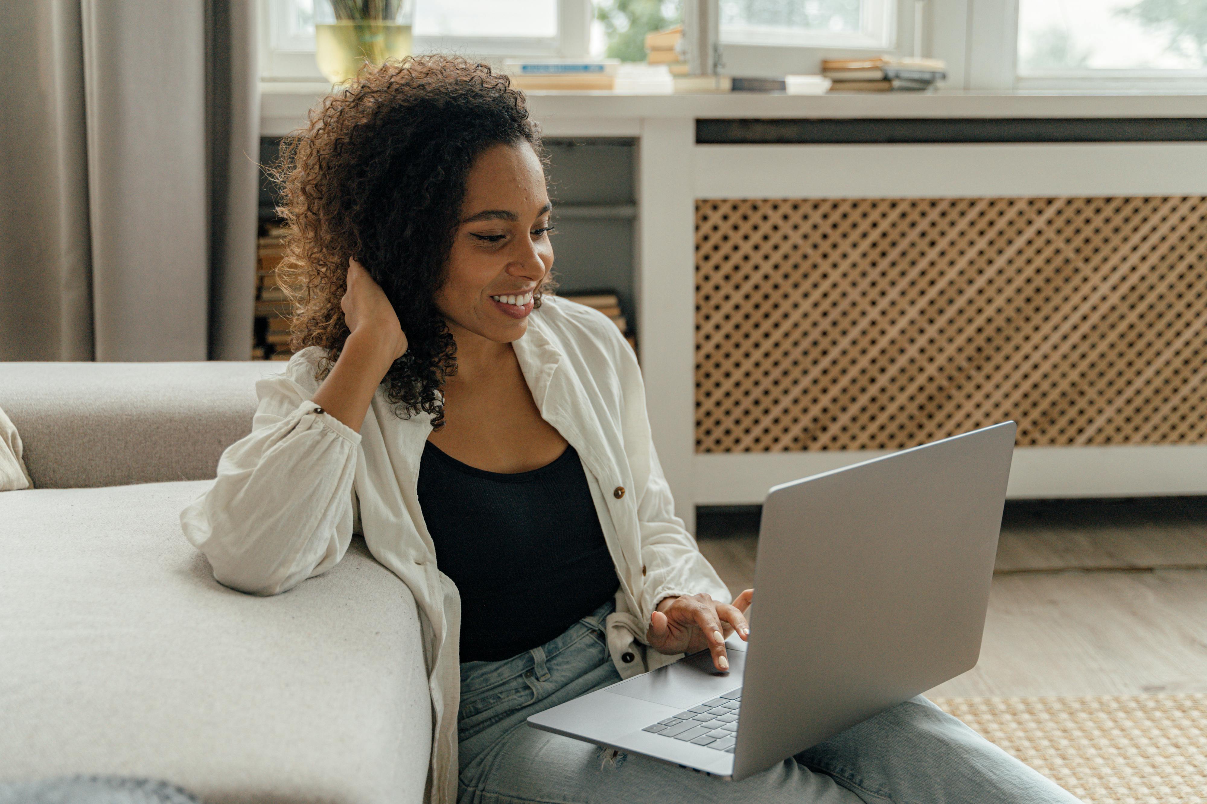 A smiling woman with curly hair sits on the floor leaning against a sofa, working comfortably on a laptop in a bright room. This relaxed yet productive scene represents the diverse and flexible work environments that top IT recruiting firms cater to.
