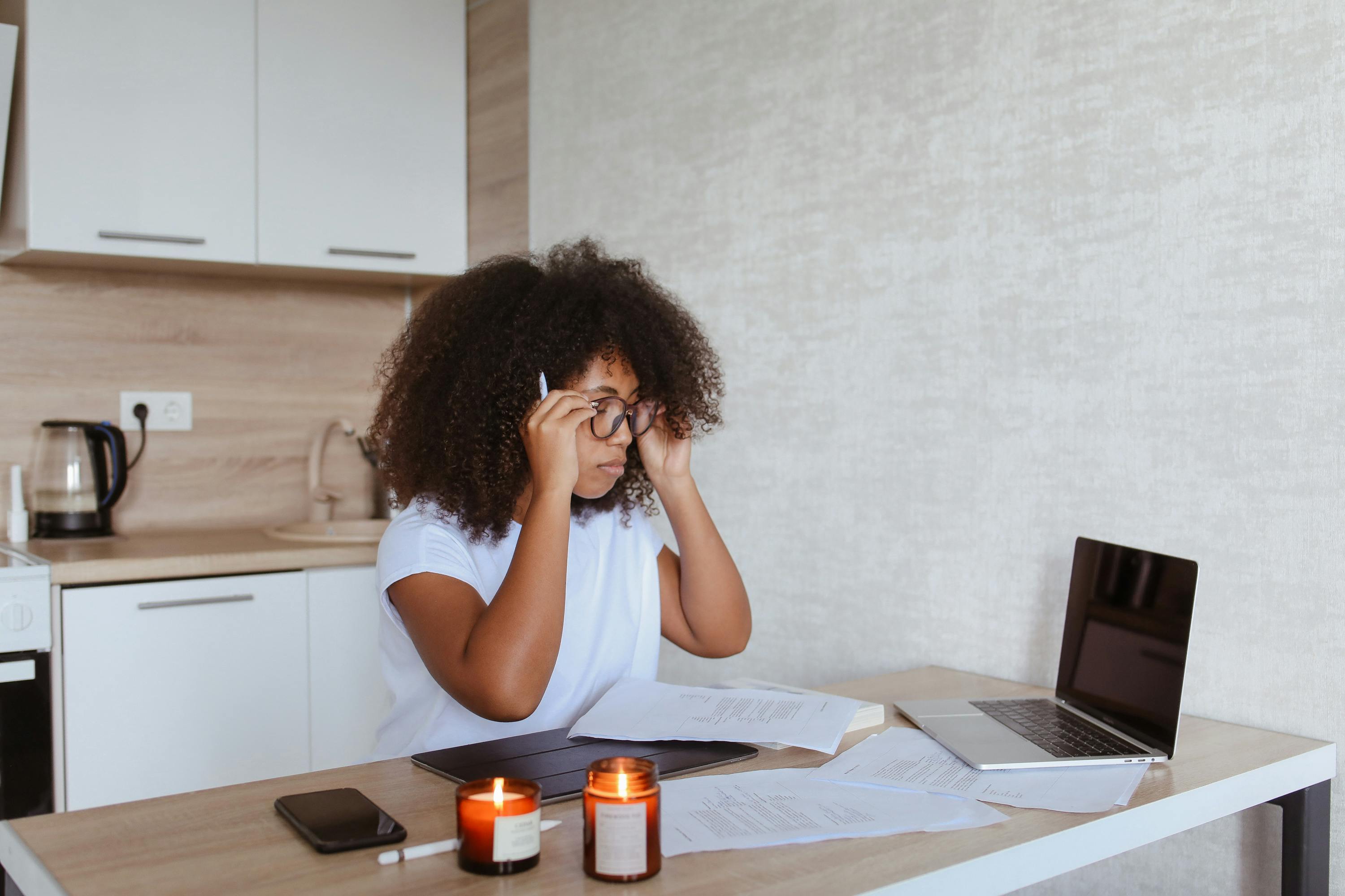 A focused woman with curly hair adjusts her glasses while working at a table with a laptop, papers, a tablet, and two lit candles. A kitchen area is visible in the background, showing a versatile workspace, reflecting the adaptable nature of roles sourced by top temp agencies.
