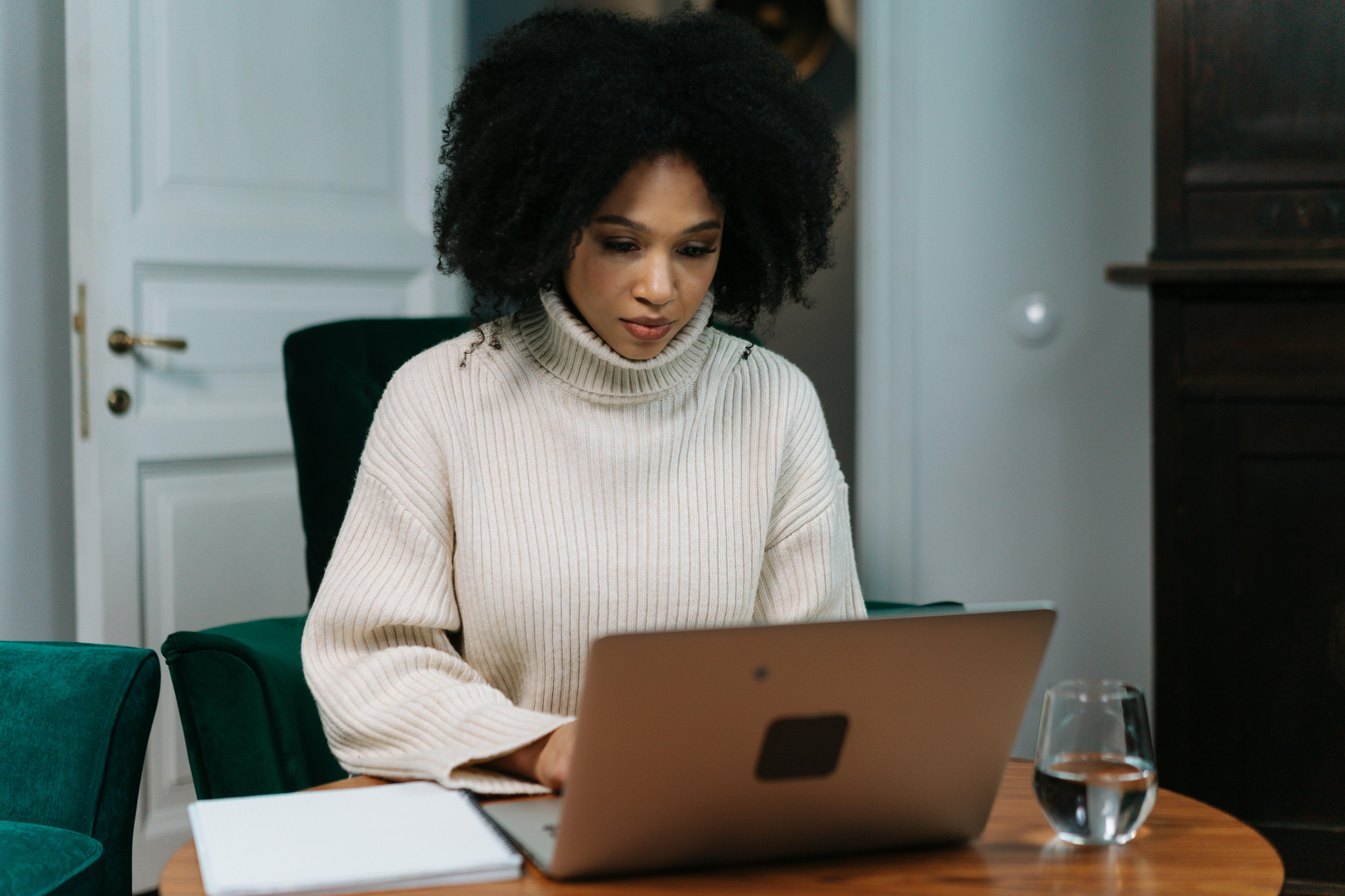 A focused woman with curly hair, wearing a cream-colored turtleneck sweater, works diligently on a laptop at a round wooden table. A glass of water and a white notebook are also on the table, reflecting the dedicated talent provided by top staff augmentation companies.