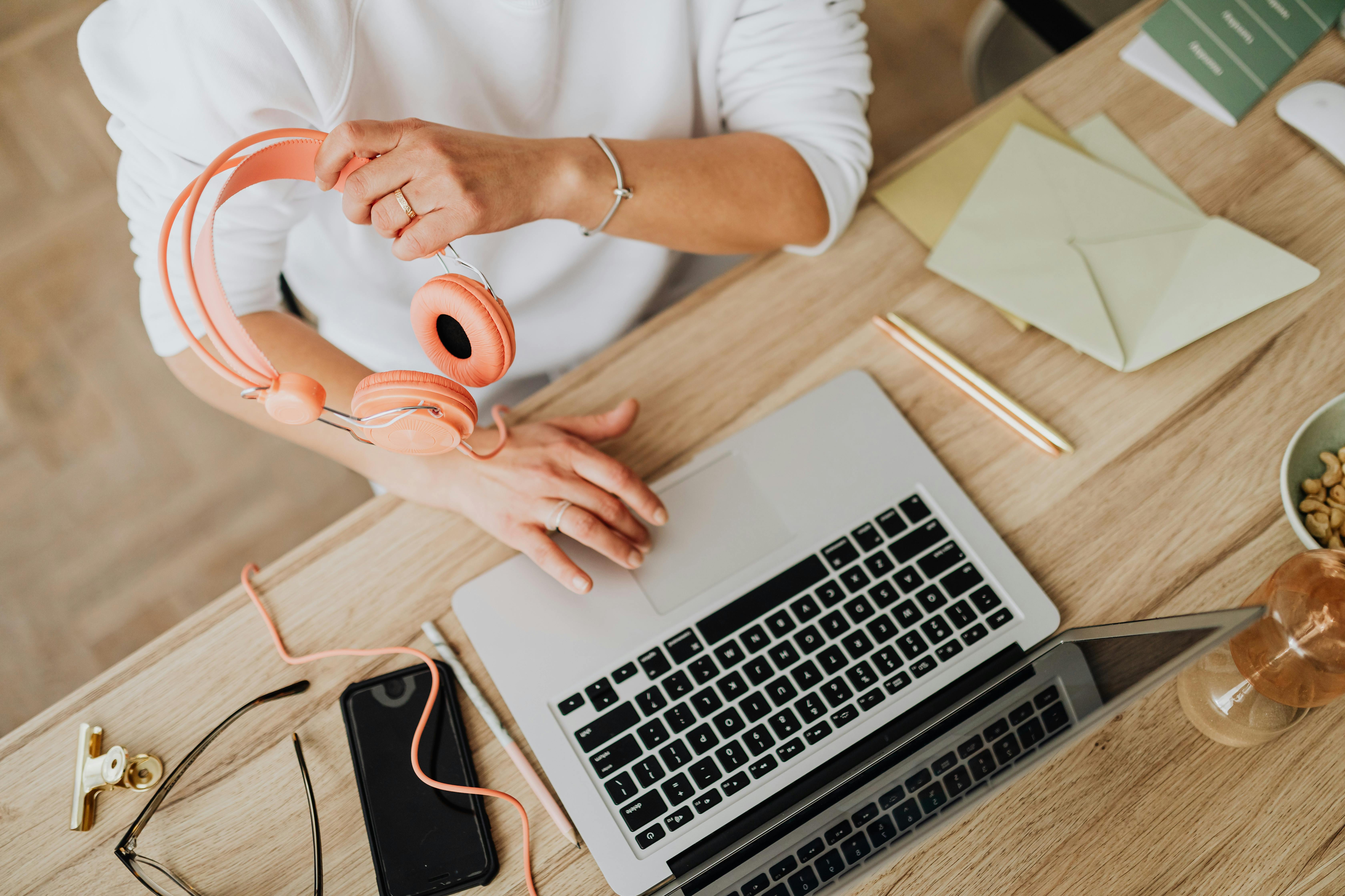An overhead shot of a person at a wooden desk, holding pink over-ear headphones, next to an open laptop, a smartphone, and various office supplies. This dynamic workspace reflects the communicative and organized nature of roles sourced by top 10 sales recruitment agencies.