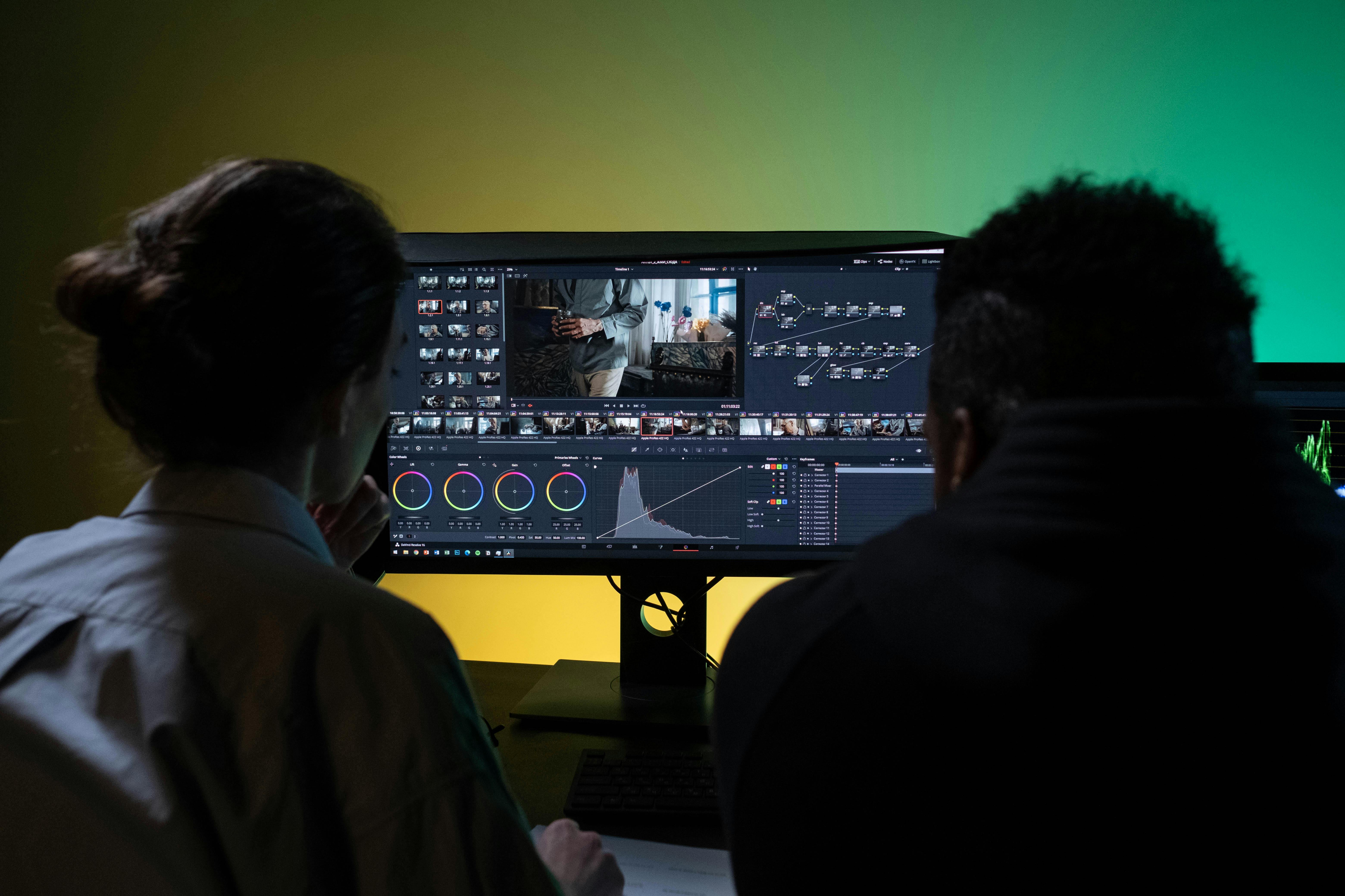 Photo of two individuals collaborating on video editing at a dual-monitor setup. Software interface shows color wheels, timeline, node-based editing, and waveform scopes. Ambient lighting in green-yellow tones, headphones, coffee mug, and a focused team environment. Software: DaVinci Resolve.