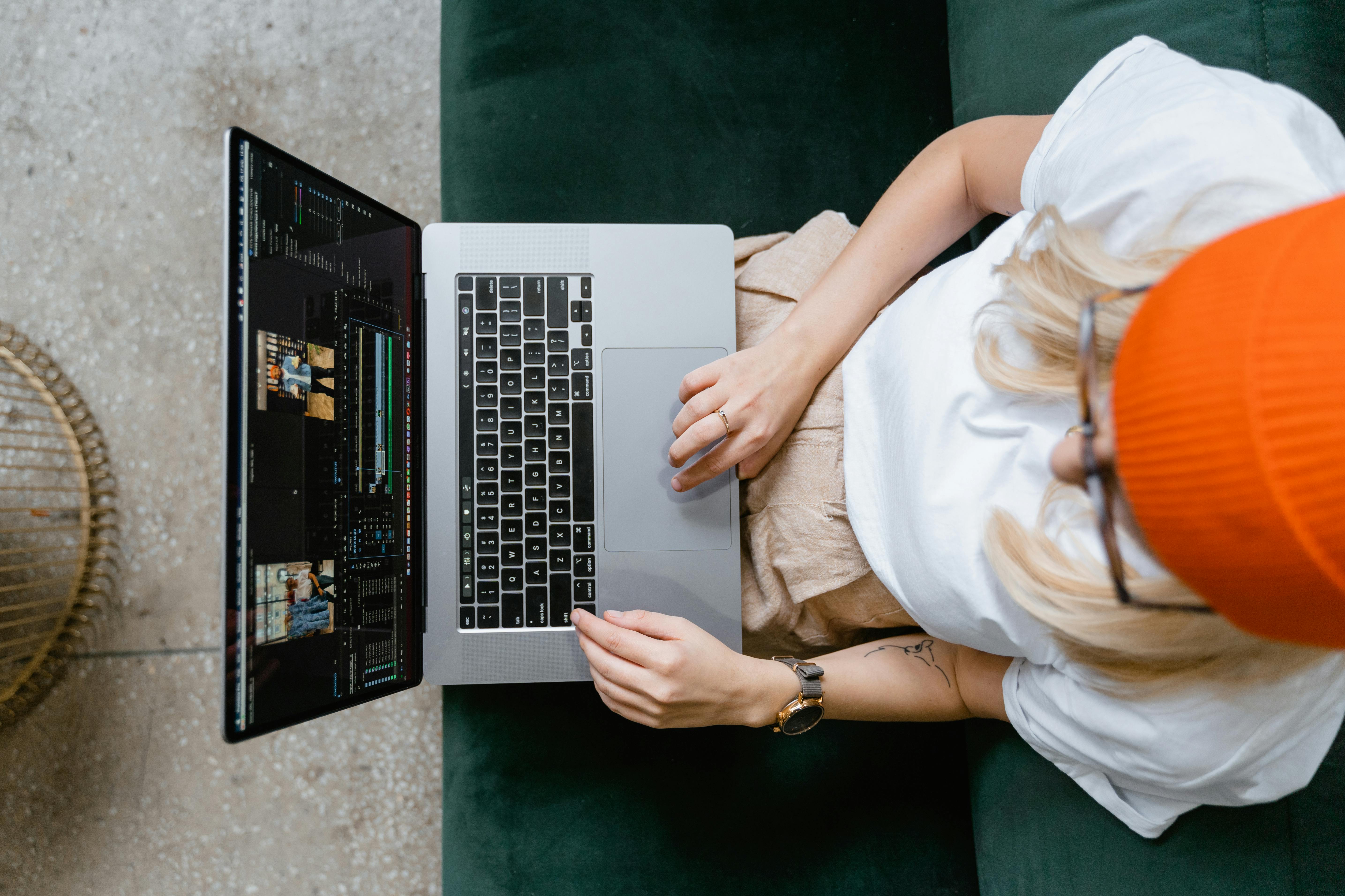 Person sitting on green couch editing video on a laptop. Wearing white t-shirt, orange beanie, and glasses. Screen shows editing software with clips, tools, and timeline. Background includes woven basket and speckled floor. Captures digital content creation in a relaxed, efficient workspace.