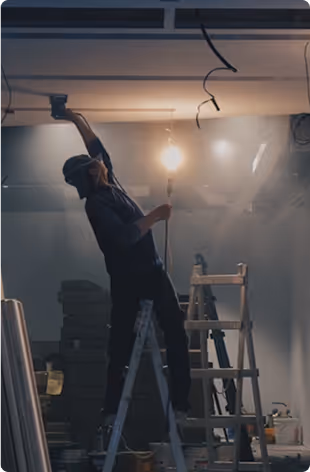 A person stands on a ladder installing a ceiling light in a dimly lit room.