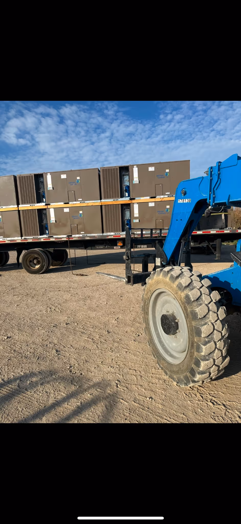 A blue forklift is lifting a pallet onto a flatbed truck loaded with large boxes.