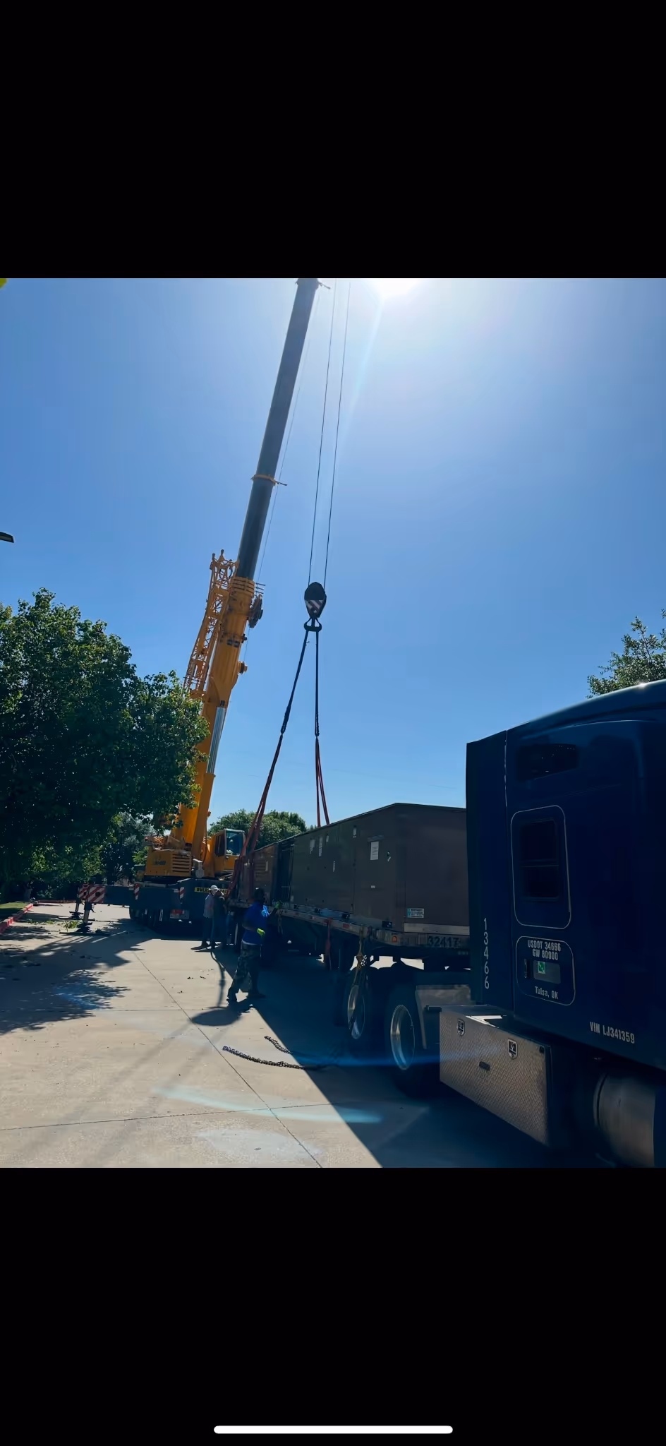 Crane lifting large container from truck on a sunny day, with workers standing nearby.