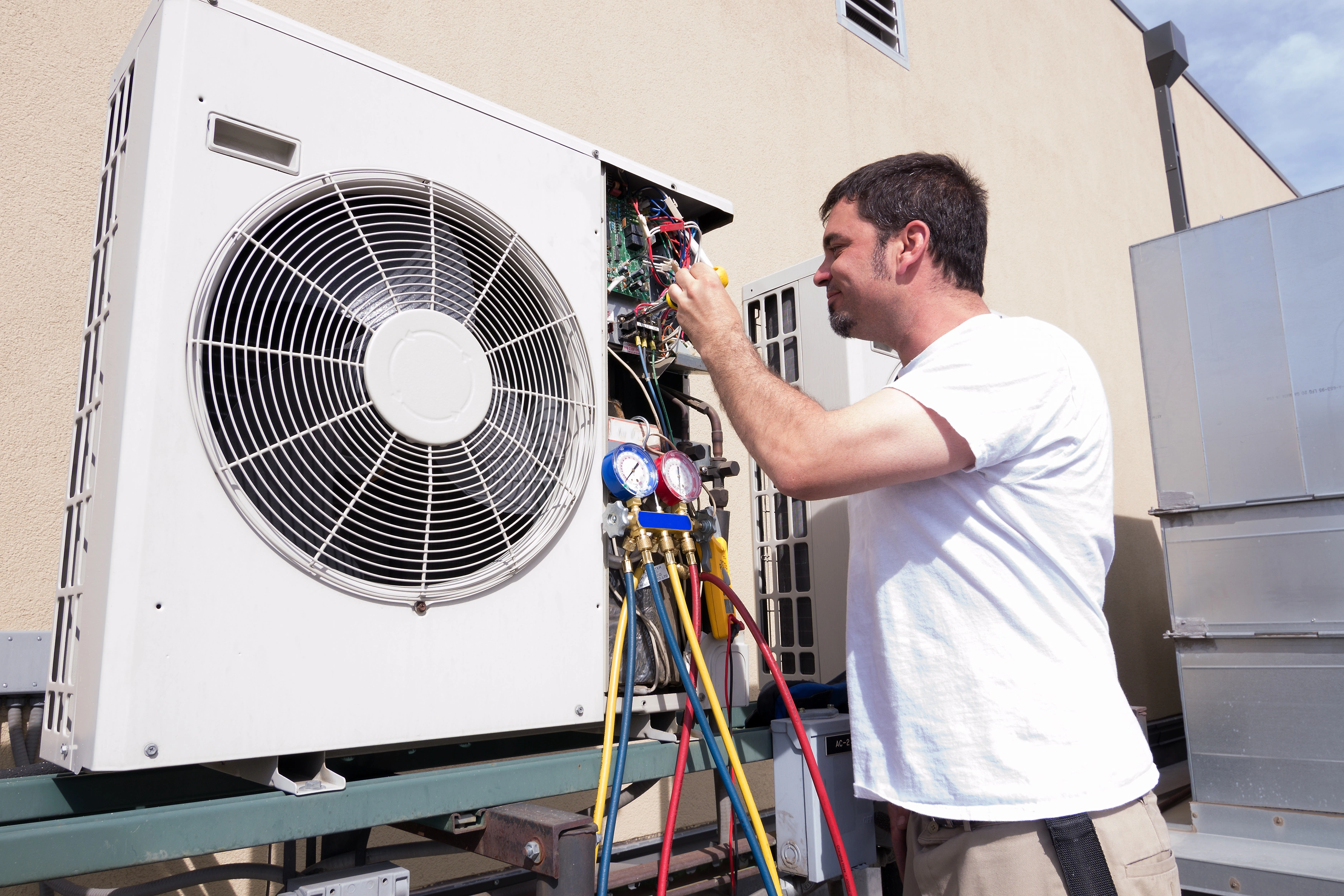 A technician in a white shirt works on an outdoor air conditioning unit. 