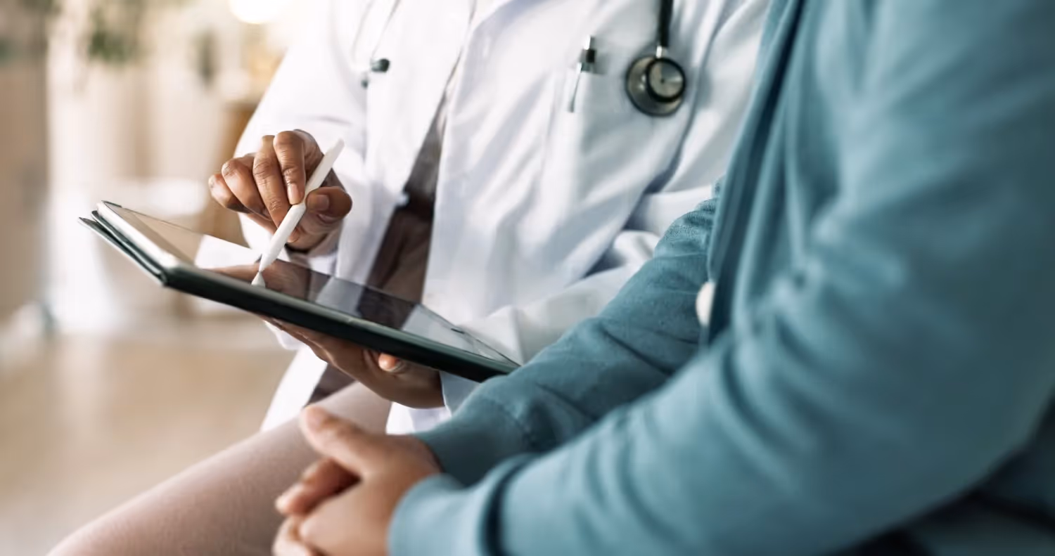 A doctor in a white coat uses a stylus to show a patient information on a digital tablet.