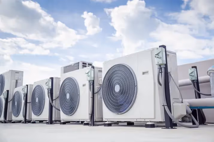 Row of white commercial air conditioning units on a rooftop under a bright blue, cloudy sky.