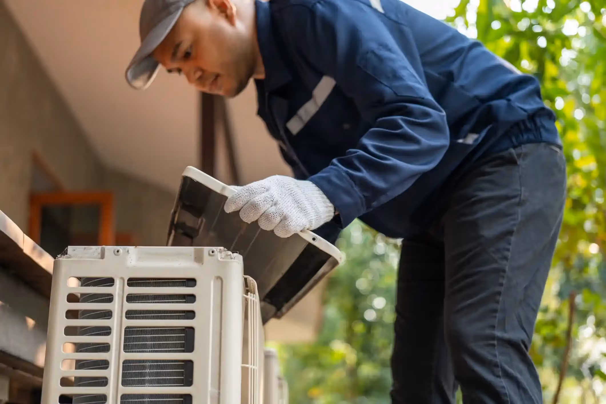 A technician in a cap and blue uniform is repairing an outdoor air conditioner unit.