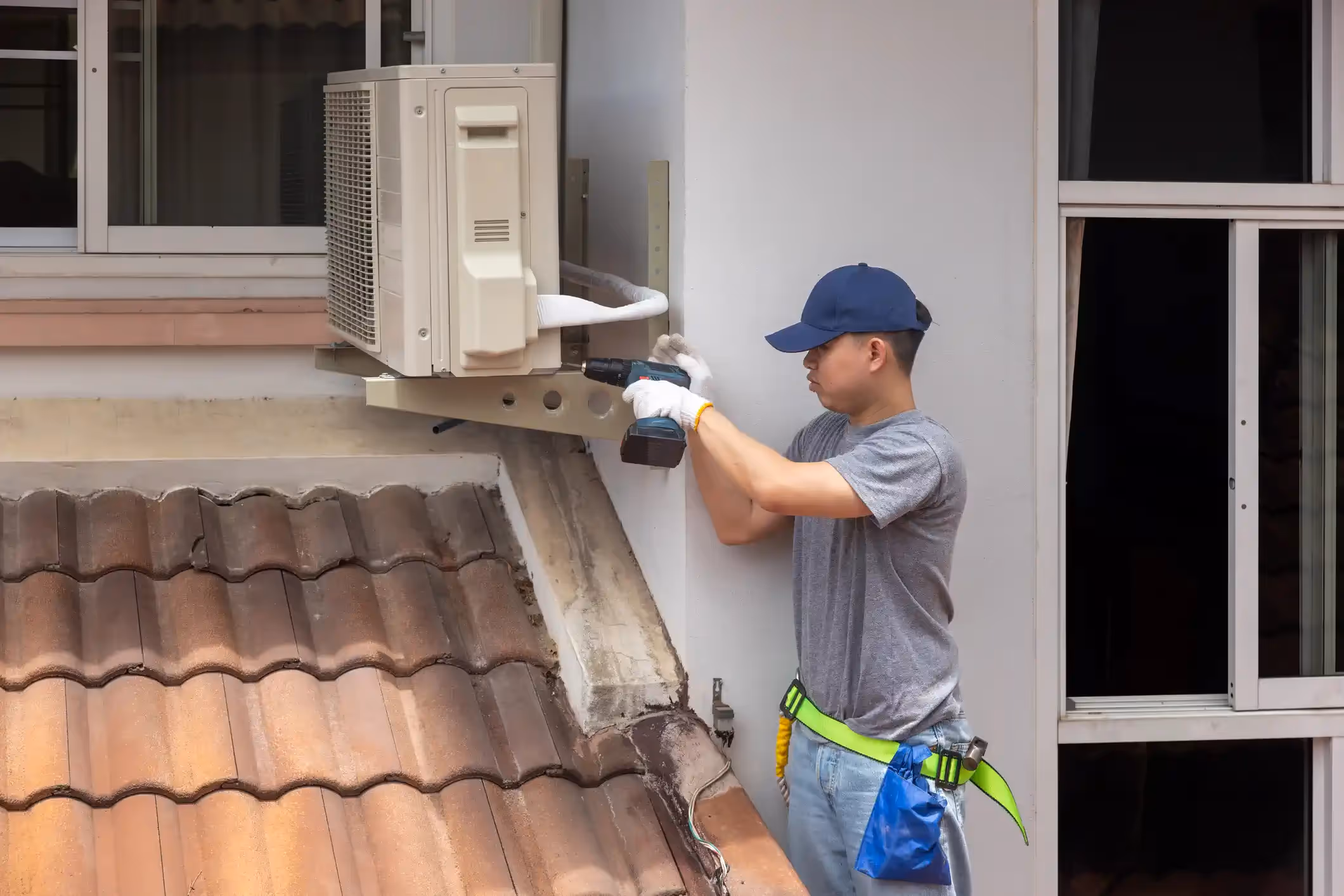  A technician, wearing a grey T-shirt, blue cap, and tool belt, is using a power drill to secure or work on the outdoor condenser unit of a mini-split AC, which is mounted on a bracket above a tiled roof.