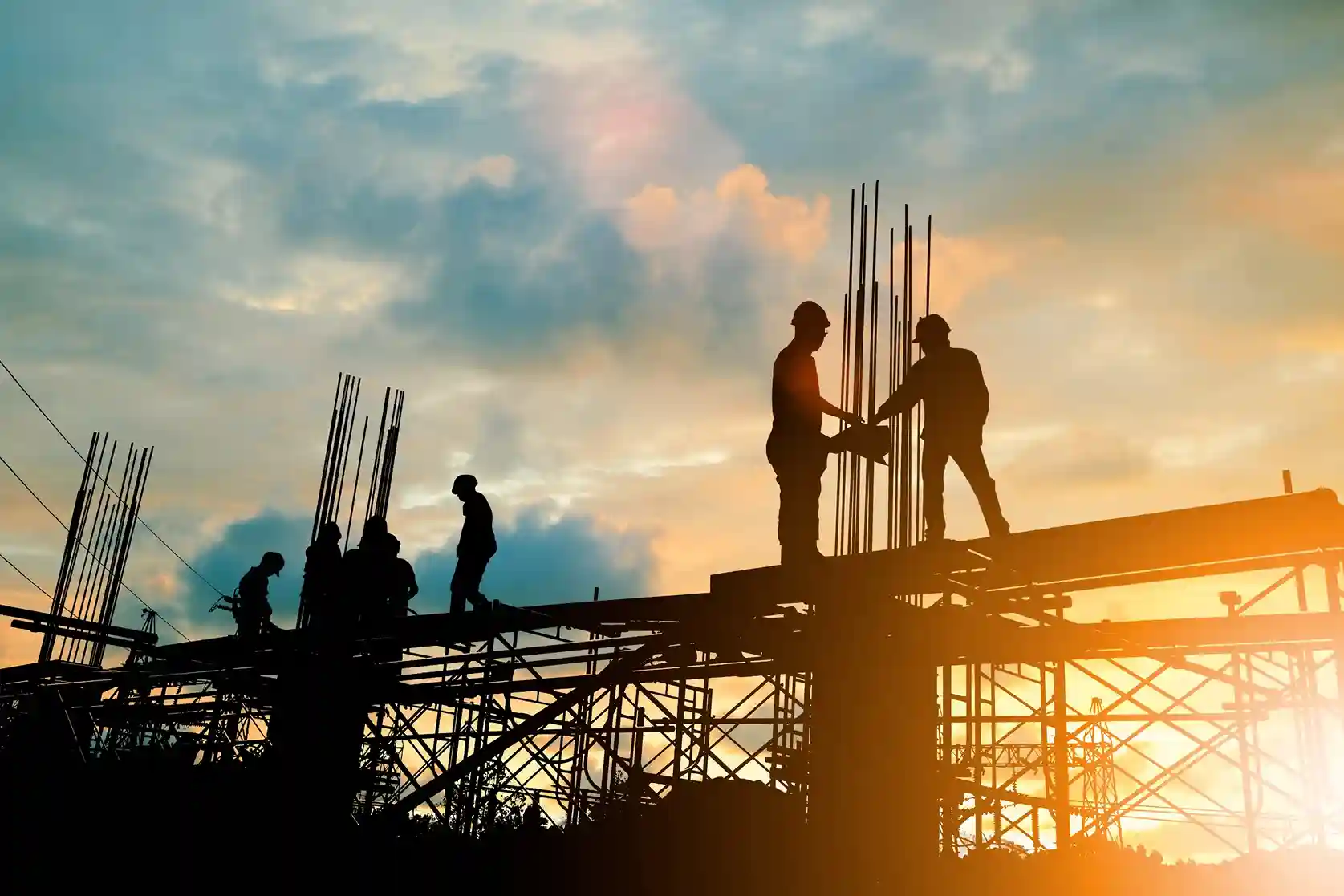Construction workers in hard hats stand on a steel beam reviewing plans during the framing phase of a commercial project.