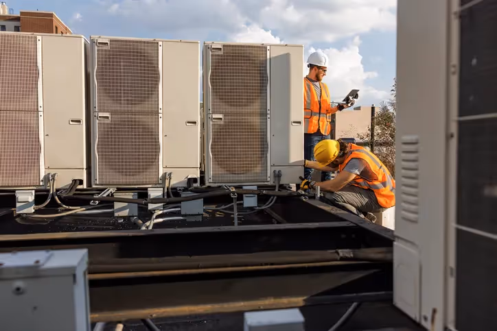Two technicians in hard hats and safety vests are working on multiple commercial AC units on a rooftop.