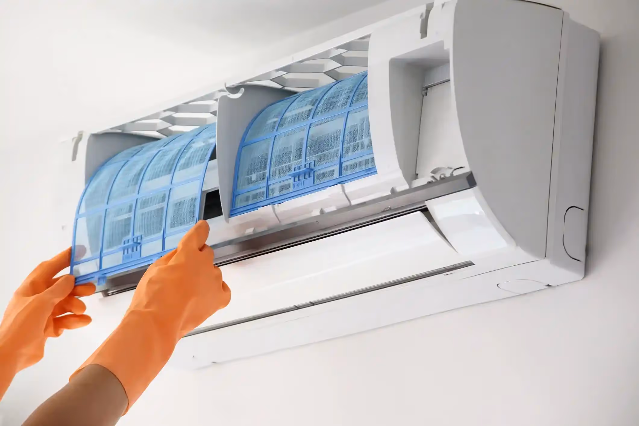 A person with orange gloves is inserting a clean, blue filter into a wall-mounted air conditioner.