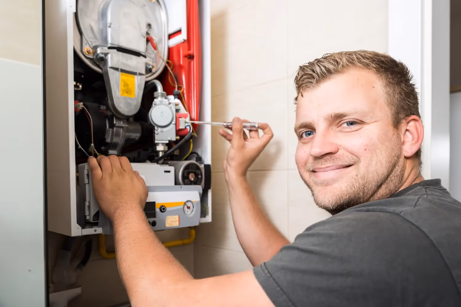 A close-up of a smiling male technician in a gray shirt, facing the camera while servicing a wall-mounted boiler or water heater. The unit's front panel is open, revealing red casing and internal components, and the technician is holding a screwdriver in his right hand.