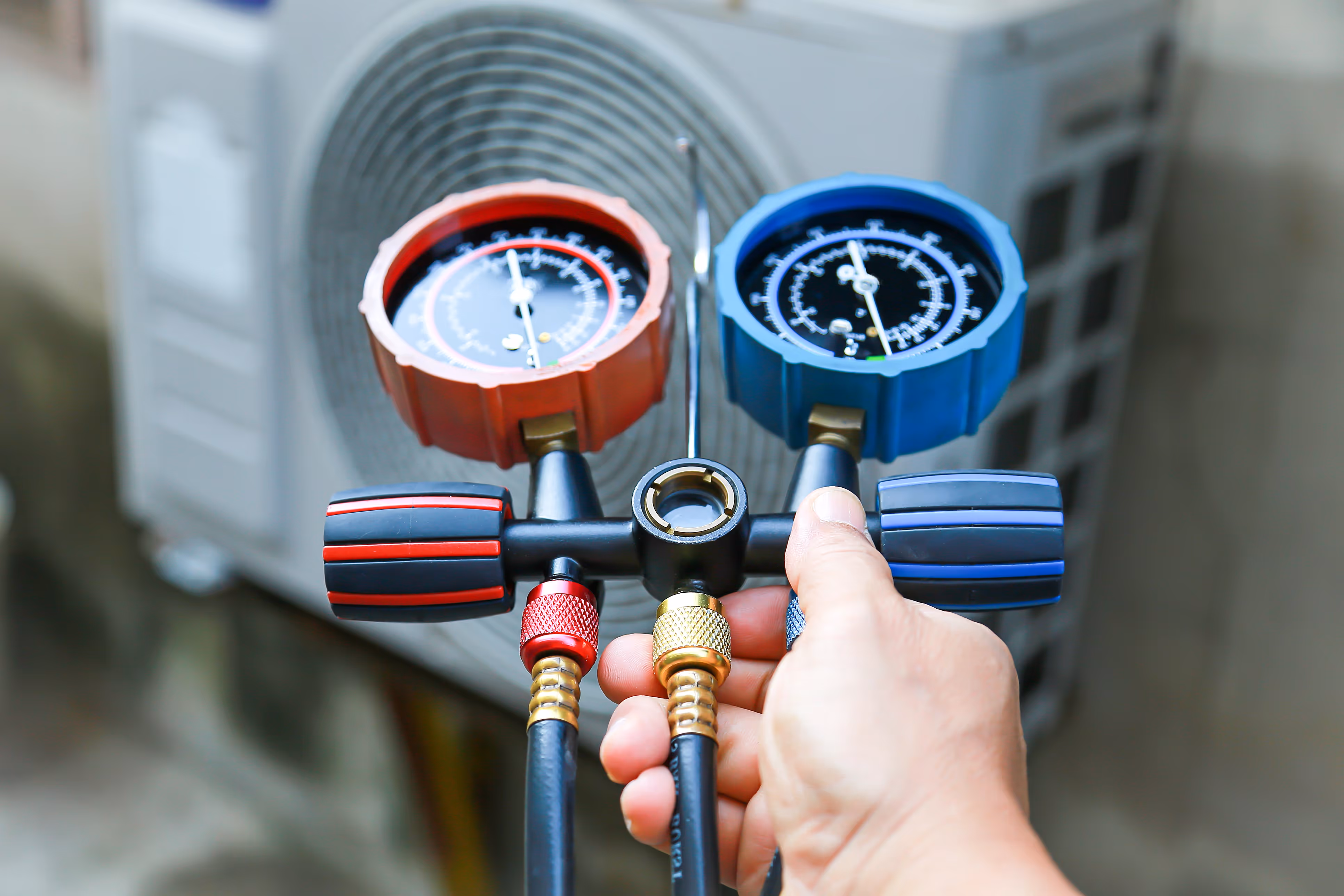Close-up of a hand holding a manifold gauge with red and blue dials near an air conditioning unit. 