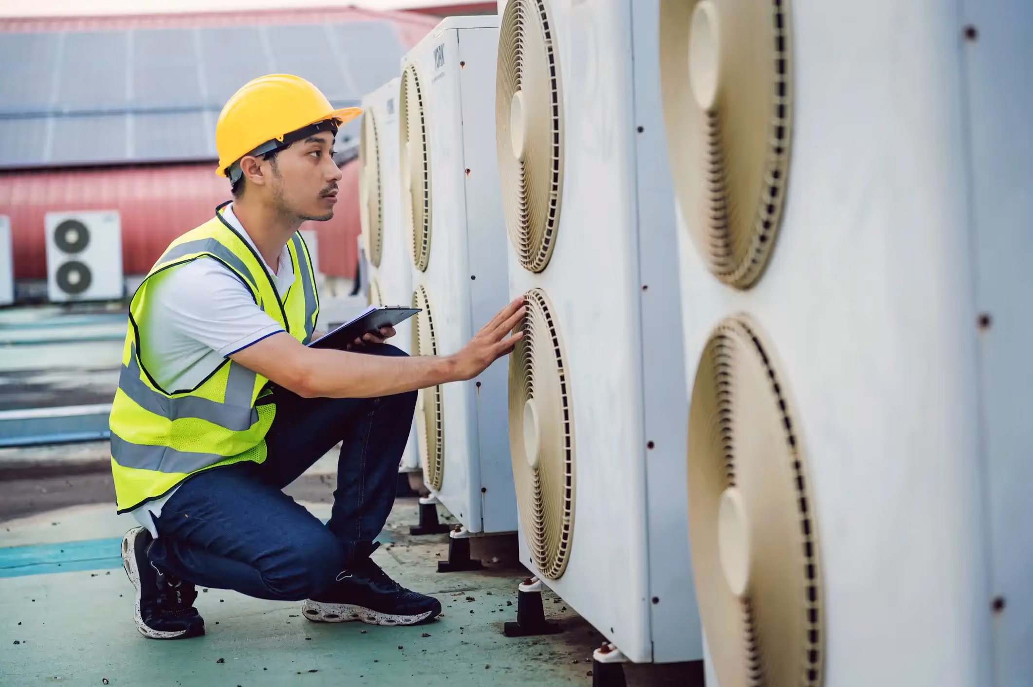  A male HVAC technician in a yellow hard hat and a lime-green high-visibility vest is kneeling on a rooftop. He is inspecting the fan units of several large, white, commercial air conditioning condenser units lined up in a row. He is holding a clipboard in his left hand and touching one of the units with his right hand.
