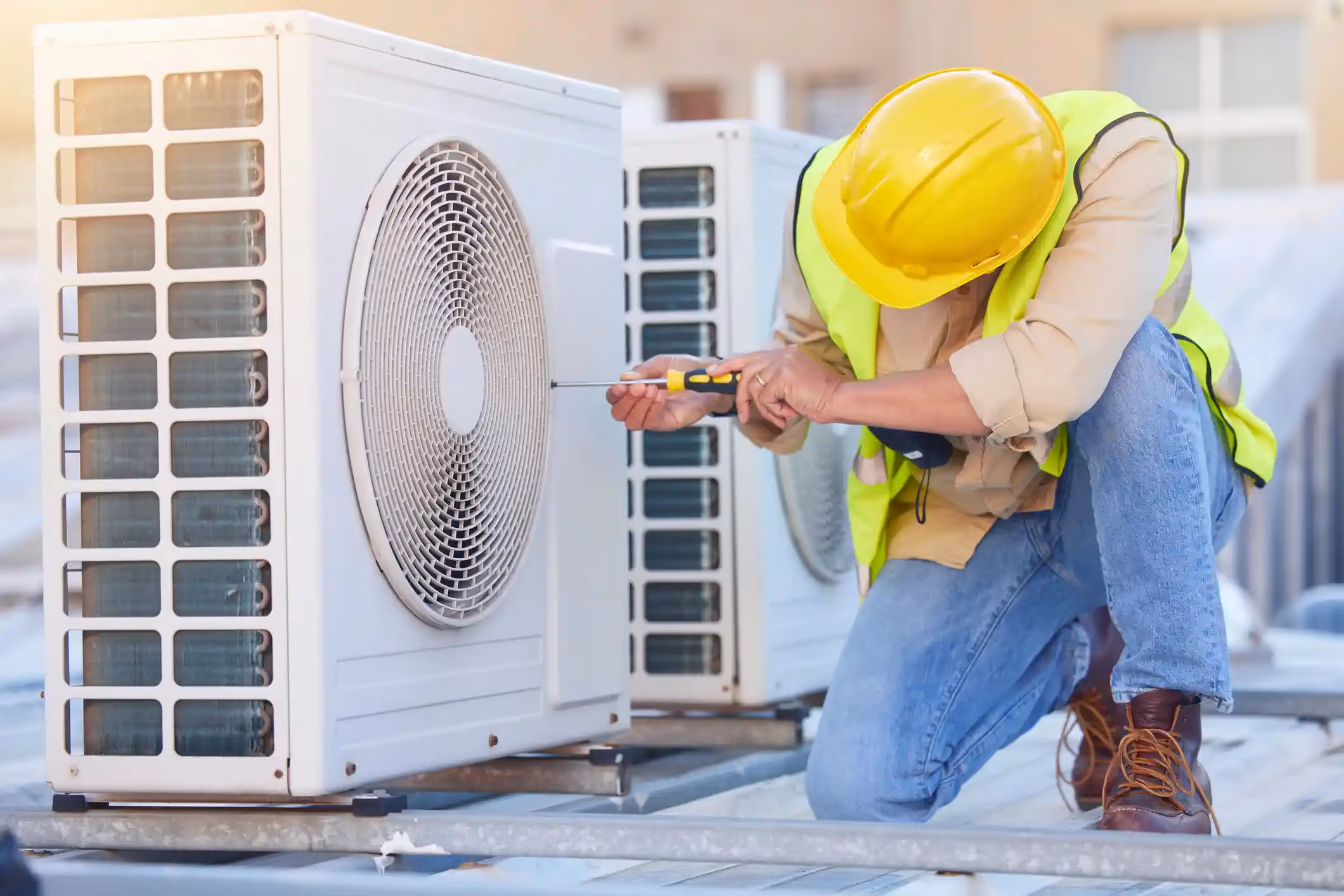 A male HVAC technician is kneeling on a rooftop, actively working on a commercial air conditioning unit. He is wearing a yellow hard hat and a lime-green high-visibility vest over a khaki shirt and jeans. He is using a screwdriver to adjust or repair the white condenser unit. The sun is shining brightly from the left, creating a lens flare and strong backlight.