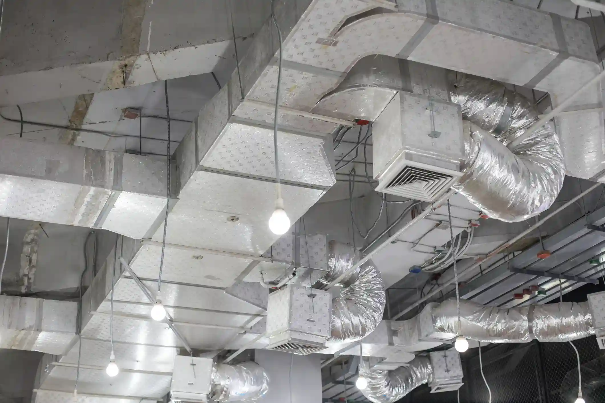  A view of a complex network of exposed HVAC ductwork suspended from a high ceiling, typical of a commercial or industrial building under construction or with an industrial design aesthetic. The ducts are a mix of large rectangular, rigid ducts and flexible round ducts, both covered in a silver, patterned insulation material or foil. Square air diffusers with grilles are attached to the rectangular ducts. Bare light bulbs hang down from wires among the ducts.