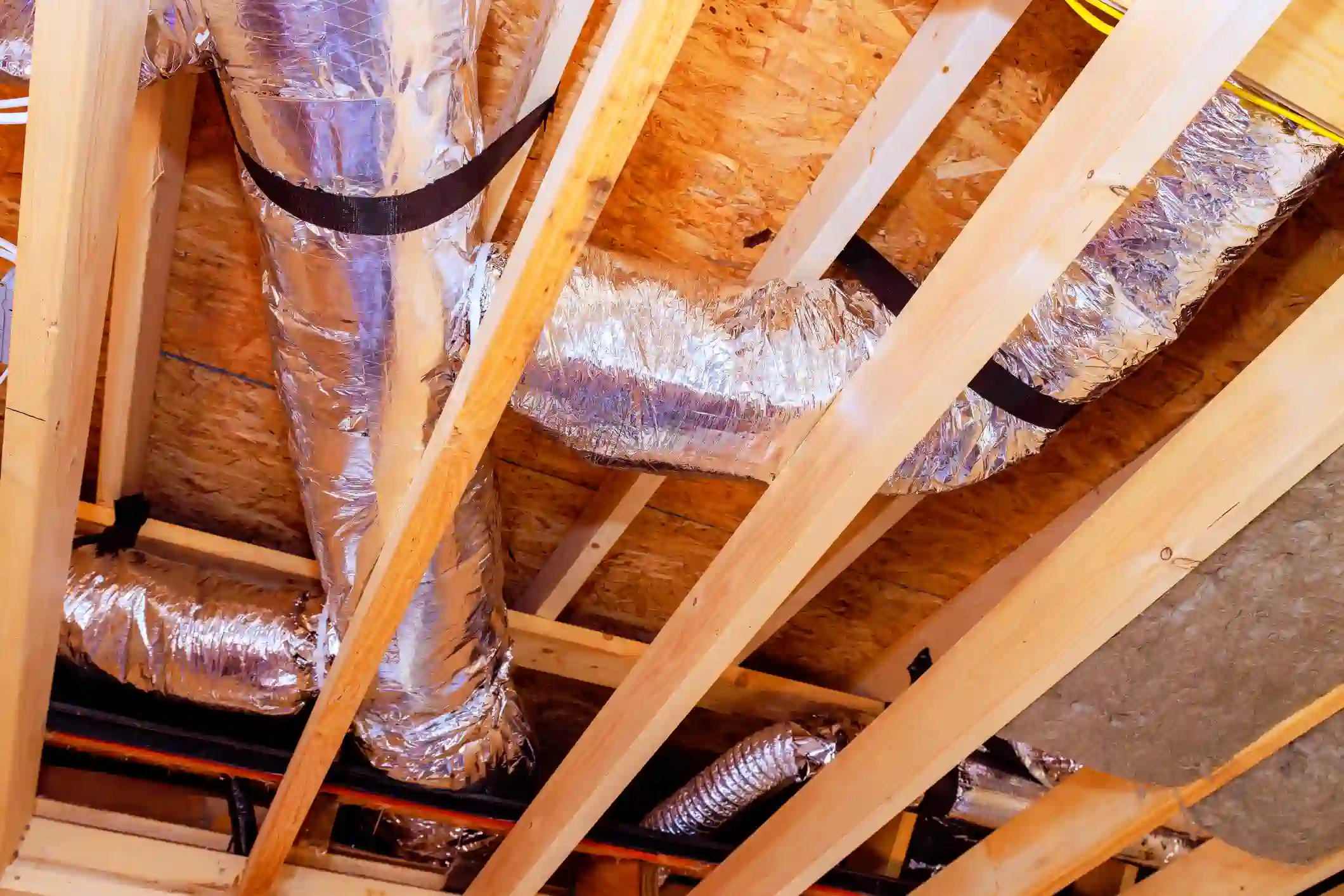 A photograph looking up into the exposed framing and sub-flooring (OSB) of a ceiling or attic during construction. Several sections of large, flexible insulated ductwork wrapped in reflective silver material are installed between the wooden joists. The ducts are secured with black strapping. Electrical wiring is also visible running through the structure.
