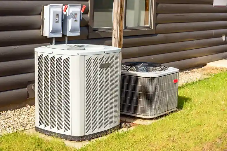Two large, gray outdoor HVAC units are side-by-side on a bed of gravel and grass next to a log cabin-style house.
