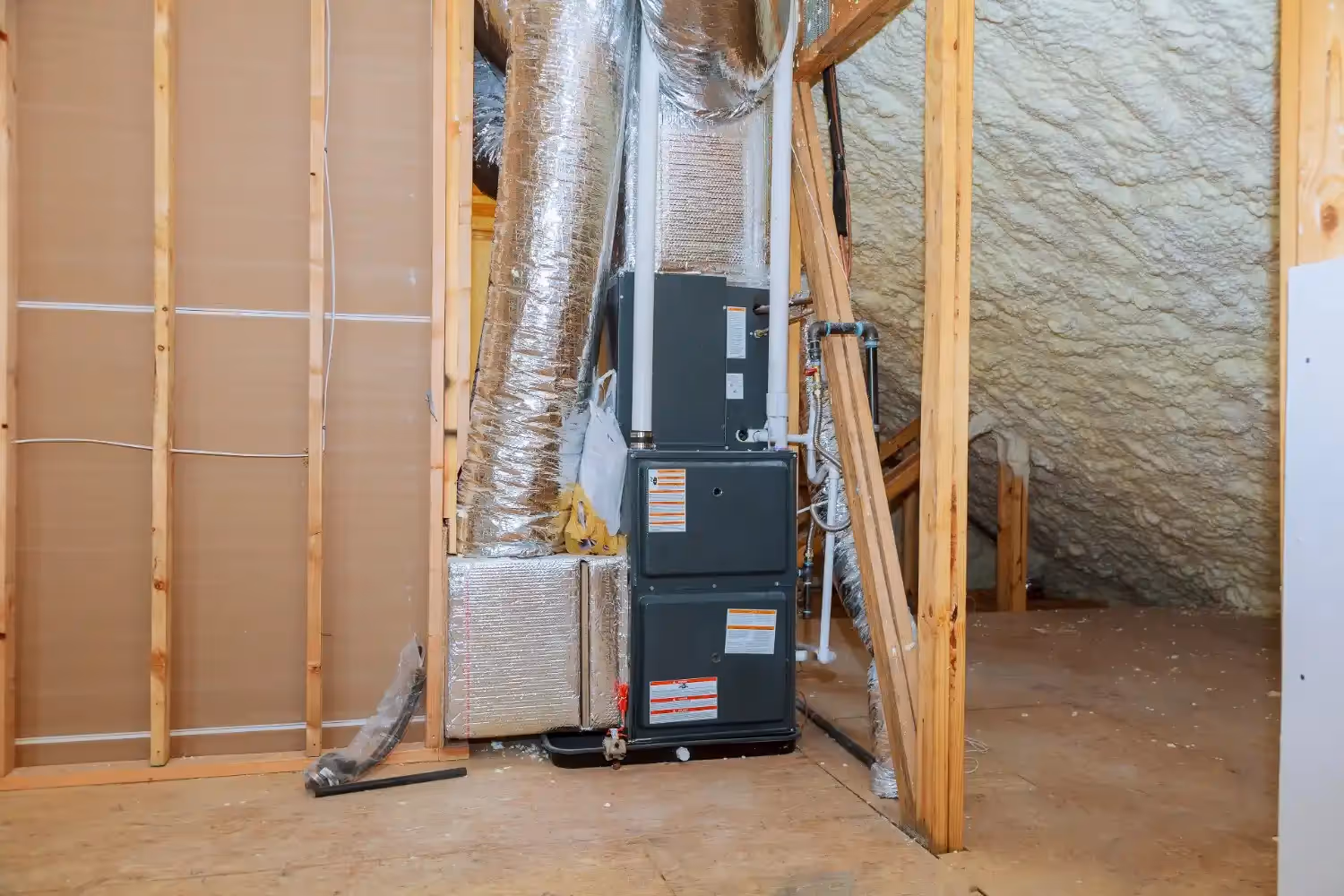 A gray, vertically stacked HVAC furnace or air handler unit is installed in an unfinished attic space. The unit is surrounded by exposed wooden framing and large, insulated flexible silver ductwork. To the left, a silver-faced insulation box is attached, and the attic walls are insulated with spray foam insulation. PVC piping is also visible for condensate drainage and exhaust.