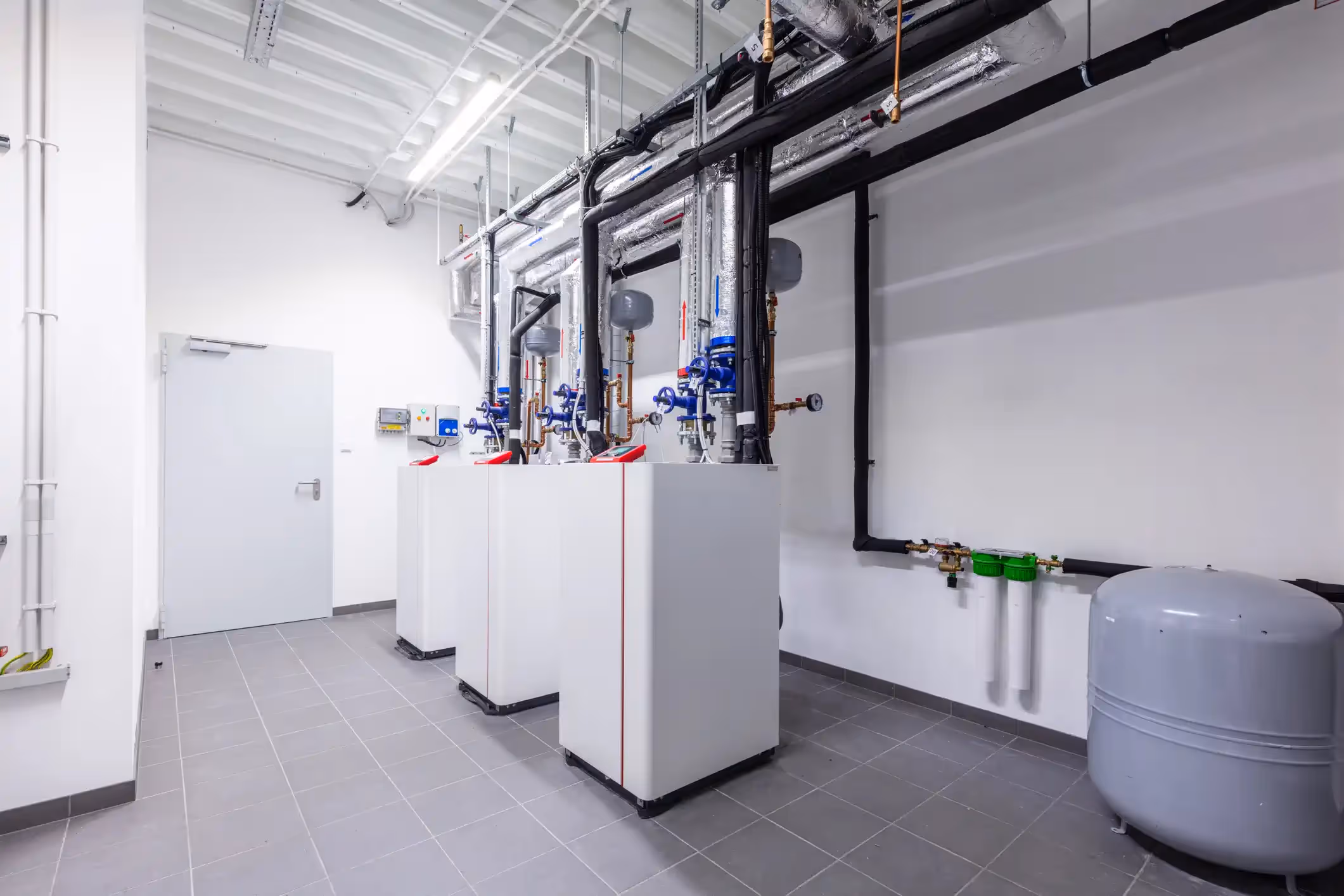 Three modern, white commercial boiler units or heat pumps are neatly lined up in a clean, white utility room with a gray tiled floor. The units are connected to a network of black-insulated pipes, manifolds with blue valves, and gauges running along the ceiling and wall. A large, gray expansion tank sits on the floor to the right, and water filtration components are visible on the wall above it. The room has bright fluorescent lighting and a white metal exit door.