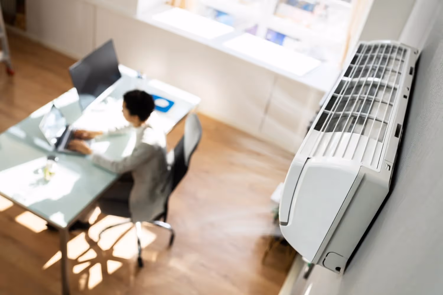  A white ductless mini-split indoor air handler unit is prominently mounted on a light-colored wall, angled toward the viewer in a modern office setting. The front panel of the unit is open, possibly for filter cleaning or service. In the blurred background, a person is seated at a white glass desk, working on a laptop near a large, bright window.