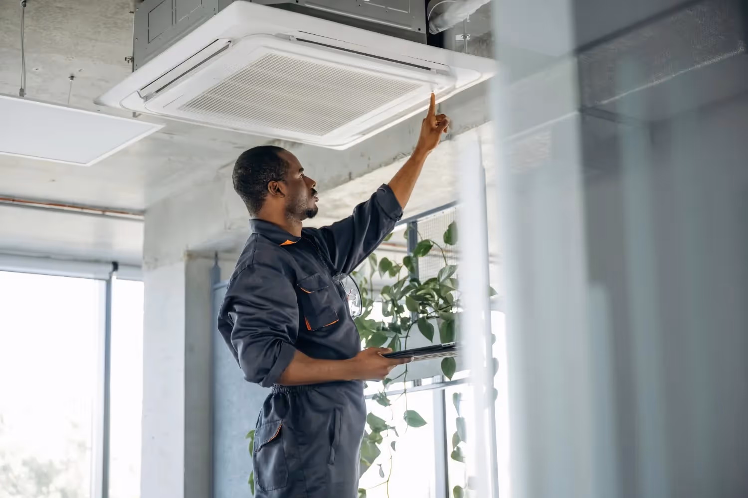  An African American technician in a dark grey uniform is standing, pointing to and inspecting a square cassette-style air conditioning unit recessed into the ceiling of a modern office space, while holding a tablet. Four HVAC professionals are then shown installing a large outdoor condenser unit on a mounting bracket against the beige siding of a house. Finally, a dark-haired technician in a black shirt is seen on a ladder, using a screwdriver to work on the bottom panel of a wall-mounted outdoor AC unit under a clear blue sky.